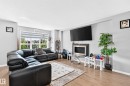 Bright living area featuring wood-finish flooring, a large window with blinds, and a built-in wall-mounted fireplace with a dark surround and stainless steel trim - 5280 21 Avenue Sw, Edmonton, AB  - Indoor Photo Showing Living Room With Fireplace 