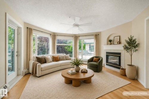 Bright living area featuring lighter tone laminate flooring, natural gas fireplace with a white mantel and arched alcove, a ceiling fan with integrated lighting, and a kitchen pass-through - #206, 9905 112 St Nw, Edmonton, AB - Indoor Photo Showing Living Room With Fireplace