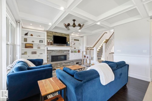 Living area featuring a stone-clad fireplace, coffered ceiling, built-in shelving, dark wood-finish flooring, and a staircase with white balusters and a wood handrail - 3 Garneau Gate, Spruce Grove, AB - Indoor Photo Showing Living Room With Fireplace