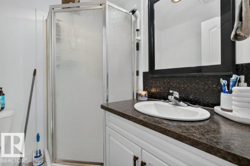 Bathroom featuring a glass-enclosed shower, white vanity cabinetry with dark hardware, a dark patterned backsplash, a countertop with an integrated sink, and a framed mirror - 12219 89 Street Nw, Edmonton, AB - Indoor Photo Showing Bathroom