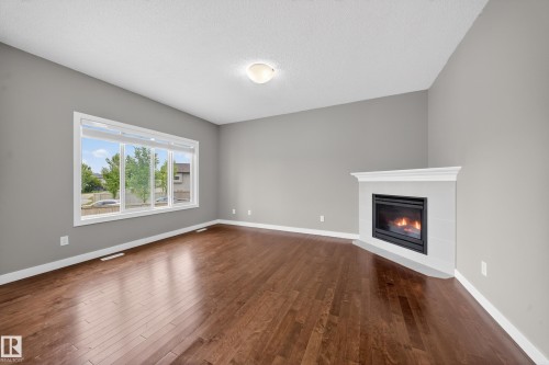 Spacious living area featuring rich wood-finish flooring, a corner gas fireplace with a white mantel, and a large multi-pane window - 3639 15A Street Nw, Edmonton, AB - Indoor Photo Showing Living Room With Fireplace