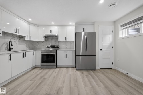 Modern kitchen featuring white cabinetry with matte black hardware, stainless steel appliances, a marble-look backsplash, light wood-finish flooring, and recessed lighting - 3639 15A Street Nw, Edmonton, AB - Indoor Photo Showing Kitchen
