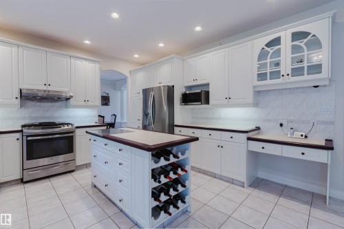 White kitchen cabinetry with stainless steel appliances, a central island featuring a built-in wine rack, light-toned tile flooring, and recessed lighting - 928 Heacock Road Nw, Edmonton, AB - Indoor Photo Showing Kitchen