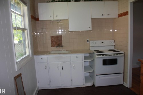 Compact kitchen featuring white cabinetry with silver hardware, a white electric range, and light-toned tiled walls with decorative accents - 5227 51 Street, Redwater, AB - Indoor Photo Showing Kitchen