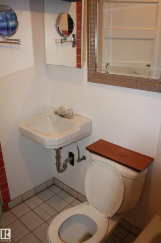 Wall-mounted sink with exposed plumbing, white toilet featuring a wood-finish tank lid, and white square floor tiles - 5227 51 Street, Redwater, AB - Indoor Photo Showing Bathroom