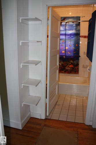 Hardwood flooring extending into the doorway of a bathroom featuring white square floor tiles and a white bathtub - 5227 51 Street, Redwater, AB - Indoor