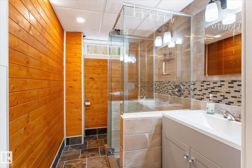 Bathroom featuring wood-finish paneling, slate-style floor tiling, a glass-enclosed shower, a built-in vanity with a single sink, and a tiled shower surround with mosaic accents - 4921 47 Avenue, Legal, AB - Indoor Photo Showing Bathroom
