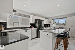 Modern kitchen featuring an island with seating, dark countertops, white cabinetry, a black tile backsplash, and stainless steel appliances - 
