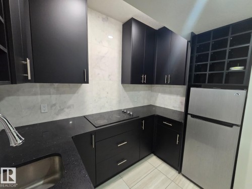 Contemporary kitchen featuring dark cabinetry with brushed nickel hardware, a black solid surface countertop, an integrated electric cooktop, and a stainless steel refrigerator - 228 36 Street Sw, Edmonton, AB - Indoor Photo Showing Kitchen