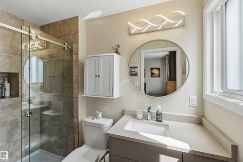 Bathroom featuring a glass-enclosed shower with tile surround and built-in niche, a white floating cabinet, a toilet, a vanity with an integrated sink and light-toned countertop, and a circular mirror - 11351 35A Avenue Nw, Edmonton, AB - Indoor Photo Showing Bathroom