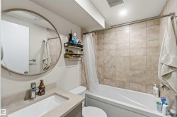Bathroom featuring a white vanity with a rectangular undermount sink, a round wall-mounted mirror, a tiled shower-over-tub with a curtain rod, and light-toned wall tiling - 