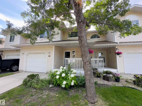 Townhome exterior featuring light beige siding, an arched window, a covered front porch with white railing, and an attached garage with a white door - #48 6608 158 Av Nw, Edmonton, AB - Outdoor With Deck Patio Veranda With Facade