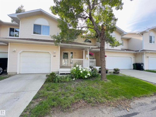 Two-story residence featuring light-colored siding, an attached garage with a concrete driveway, a covered front porch with white railings, and a landscaped garden bed - #48 6608 158 Av Nw, Edmonton, AB - Outdoor With Deck Patio Veranda With Facade