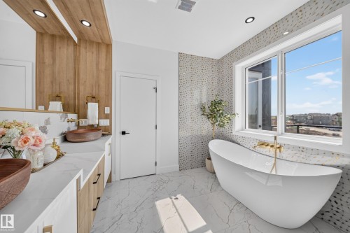 Modern bathroom featuring a freestanding soaking tub, large window, patterned tile accent wall, wood-finish vanity with dual vessel sinks, and white marble-look countertops - 423 Robinson Bv, Leduc, AB - Indoor Photo Showing Bathroom