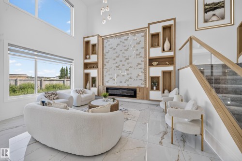 Living area featuring a floor-to-ceiling accent wall with integrated shelving and an electric fireplace, soaring ceilings, large format tile flooring, expansive windows, and a staircase with glass railing - 423 Robinson Bv, Leduc, AB - Indoor With Fireplace