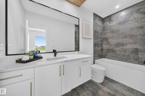 Contemporary bathroom featuring an expansive vanity with a white countertop and gold-finish hardware, a large frameless mirror, a built-in tub with dark gray tiled surround, and wood-finish flooring - 423 Robinson Bv, Leduc, AB - Indoor Photo Showing Bathroom