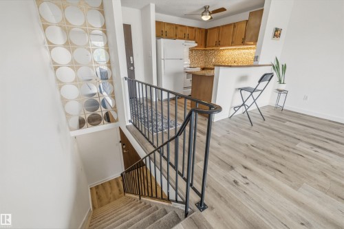Open-concept kitchen and living area featuring wood-finish flooring, a kitchen with wood cabinetry and mosaic tile backsplash, a breakfast bar, and a staircase with a carpet runner and black metal railing - #8 10931 83 St Nw, Edmonton, AB - Indoor