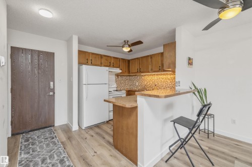 Open-concept kitchen area featuring wood-finish flooring, warm-toned cabinetry, a mosaic tile backsplash, and a breakfast bar peninsula with a light-toned countertop - #8 10931 83 St Nw, Edmonton, AB - Indoor Photo Showing Kitchen
