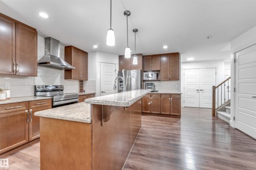 Modern kitchen featuring wood-finish flooring, stainless steel appliances, a large island with a light-toned countertop, pendant lighting, and abundant cabinetry - 6727 20 Ave Sw, Edmonton, AB - Indoor Photo Showing Kitchen With Double Sink With Upgraded Kitchen