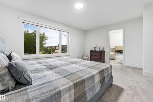 Expansive bedroom featuring light gray carpeting, a large picture window, and a white framed doorway opening to a bathroom with tiled flooring - 6727 20 Ave Sw, Edmonton, AB - Indoor Photo Showing Bedroom