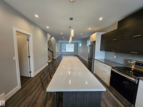 Modern kitchen featuring a large island with white countertop, dark wood-finish cabinetry, stainless steel appliances, and wood-finish flooring - 9311 66 Avenue Nw, Edmonton, AB - Indoor