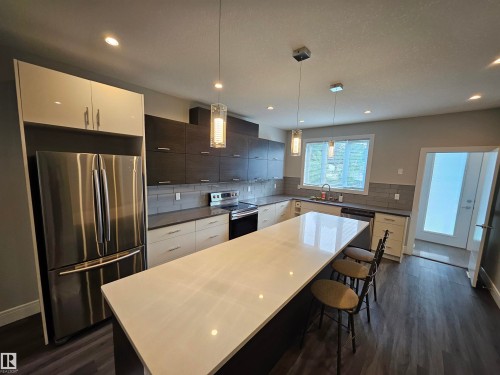 Contemporary kitchen featuring an expansive island with a light-toned countertop, a stainless steel refrigerator, dark wood-finish cabinetry, gray subway tile backsplash, and recessed lighting with pendant fixtures - 9311 66 Avenue Nw, Edmonton, AB - Indoor Photo Showing Kitchen With Upgraded Kitchen