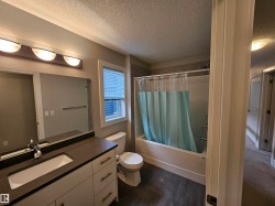 Bathroom featuring a vanity with a rectangular sink and dark countertop, a toilet, a bathtub with shower, wood-finish flooring, and a window with blinds - 