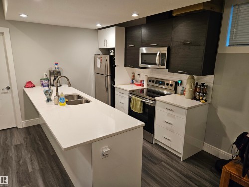 Contemporary kitchen featuring a large island with an undermount sink, stainless steel appliances, wood-finish flooring, recessed lighting, and mixed cabinetry with white uppers and dark lower cabinets - 9311 66 Avenue Nw, Edmonton, AB - Indoor Photo Showing Kitchen With Double Sink