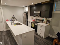 Contemporary kitchen featuring a large island with an undermount sink, stainless steel appliances, wood-finish flooring, recessed lighting, and mixed cabinetry with white uppers and dark lower cabinets - 