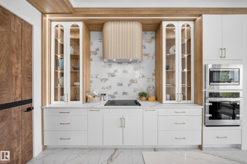Modern kitchen featuring white cabinetry with gold-tone hardware, a sleek cooktop, and a custom wood-finish range hood - 112 Ross Close, Leduc, AB - Indoor Photo Showing Kitchen