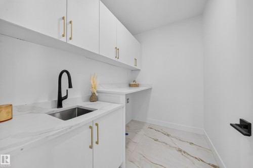 Laundry room featuring white cabinetry with gold-tone hardware, white countertops, a stainless steel sink with a matte black gooseneck faucet, and large format floor tiles with a veined pattern - 112 Ross Close, Leduc, AB - Indoor Photo Showing Other Room