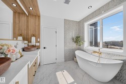 Modern bathroom featuring a freestanding soaking tub, large window, patterned tile accent wall, wood-finish vanity with dual vessel sinks, and white marble-look countertops - 