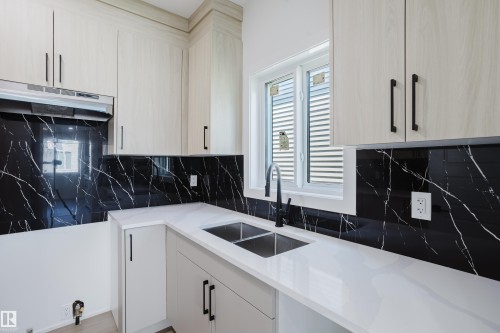 Modern kitchen featuring light wood-finish cabinetry with matte black hardware, white countertops, a deep double basin sink with a gooseneck faucet, and a dramatic black patterned backsplash - 8728 182 Avenue Nw, Edmonton, AB - Indoor Photo Showing Kitchen With Double Sink