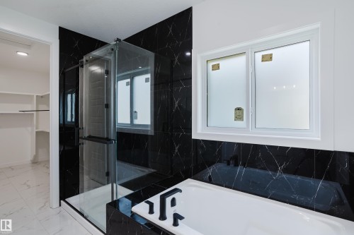 Contemporary bathroom featuring a built-in soaking tub with black fixtures, a walk-in shower with clear glass enclosure, black marble-look tile walls, white rectangular window, and white marble-look floor tile - 8728 182 Avenue Nw, Edmonton, AB - Indoor Photo Showing Bathroom