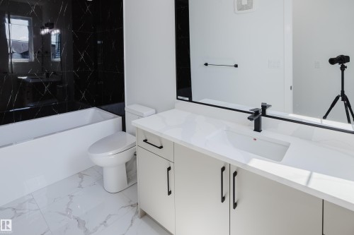 Bathroom featuring a white vanity with an integrated sink and matte black faucet, a large mirror, a white bathtub with black tiled surround, a white toilet, and white marble-look floor tiling - 8728 182 Avenue Nw, Edmonton, AB - Indoor Photo Showing Bathroom