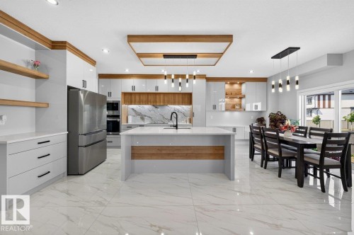 Open-concept kitchen and dining area featuring a central island, integrated stainless steel appliances, a marble-patterned backsplash, white cabinetry with wood-finish accents, and large format tile flooring - 6001 19 Street, Nisku, AB - Indoor