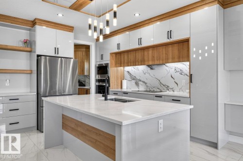 Spacious kitchen featuring an expansive central island with a built-in sink, sleek stainless steel refrigerator, integrated built-in appliances, a striking marble-look slab backsplash, and a contemporary linear chandelier - 6001 19 Street, Nisku, AB - Indoor Photo Showing Kitchen With Upgraded Kitchen