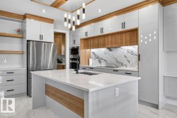 Spacious kitchen featuring an expansive central island with a built-in sink, sleek stainless steel refrigerator, integrated built-in appliances, a striking marble-look slab backsplash, and a contemporary linear chandelier - 