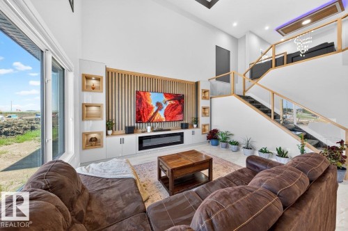 Spacious living area featuring a double-height ceiling, large windows, wood-finish accent wall with integrated shelving, built-in media console with fireplace, and a contemporary staircase with glass railing - 6001 19 Street, Nisku, AB - Indoor Photo Showing Living Room