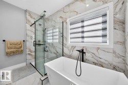 Contemporary bathroom featuring a freestanding soaking tub with a floor-mounted matte black faucet, a glass-enclosed shower with black hardware, and large format marble-look wall and floor tiles - 
