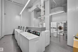 On-site laundry facility featuring multiple commercial-grade washers and dryers, suspended linear lighting, exposed overhead ductwork, and durable speckled flooring - 