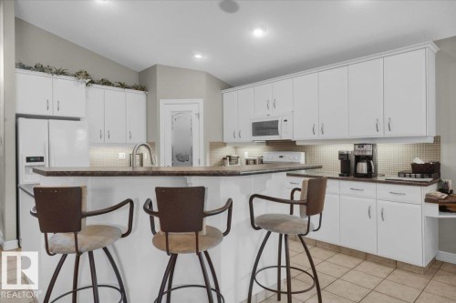 Kitchen featuring white cabinetry with silver hardware, an integrated kitchen island with counter seating, a stainless steel sink with a gooseneck faucet, a white side-by-side refrigerator, and tile flooring - 928 Thompson Place Nw, Edmonton, AB - Indoor Photo Showing Kitchen