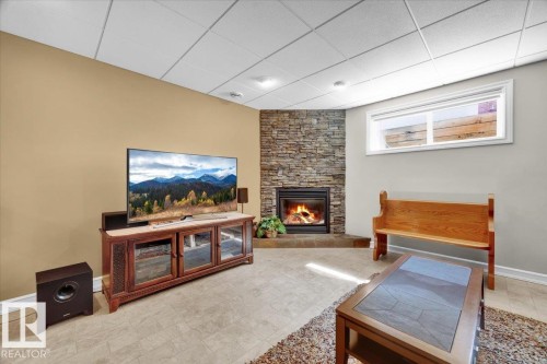 Finished basement recreation room featuring a wood-finish media console with glass-front cabinetry, gas fireplace with stone surround, recessed ceiling lighting, neutral carpet flooring, and natural light from transom windows - 928 Thompson Place Nw, Edmonton, AB - Indoor With Fireplace