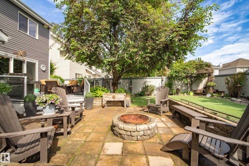 Stone paver patio featuring a built-in stone fire pit, a raised deck with glass railings, a grass lawn, mature trees, and perimeter fencing - 416 Sparling Crest Sw, Edmonton, AB - Outdoor