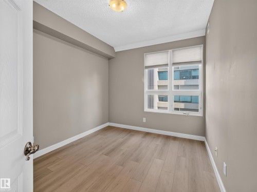 Neutral-toned room featuring a large window with horizontal blinds, wood-finish flooring, white baseboards, and crown molding - #1208 9939 109 St Nw, Edmonton, AB - Indoor Photo Showing Other Room