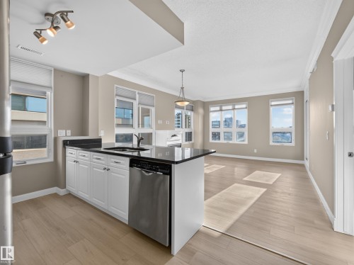 Open-concept kitchen and living area featuring white cabinetry, dark countertops, a stainless steel dishwasher, light wood-finish flooring, and multiple windows - #1208 9939 109 St Nw, Edmonton, AB - Indoor Photo Showing Kitchen With Upgraded Kitchen