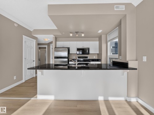 Kitchen island with black countertop, stainless steel appliances, white cabinetry, wood-finish flooring, and crown molding - #1208 9939 109 St Nw, Edmonton, AB - Indoor Photo Showing Kitchen With Double Sink