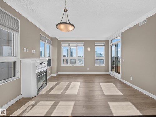 Spacious living area featuring wood-finish flooring, a decorative fireplace with white mantel, a bronze chandelier, and multiple windows including a sliding glass door - #1208 9939 109 St Nw, Edmonton, AB - Indoor Photo Showing Other Room