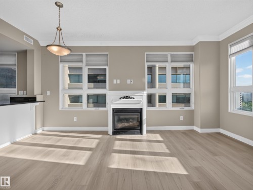Spacious living area featuring light wood-finish flooring, a decorative fireplace with a white mantel, and multiple windows with blinds - #1208 9939 109 St Nw, Edmonton, AB - Indoor With Fireplace