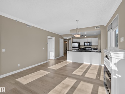Open-concept living area with wood-finish flooring, crown molding, a built-in fireplace, and a kitchen featuring white cabinetry, dark countertops, and stainless steel appliances - #1208 9939 109 St Nw, Edmonton, AB - Indoor Photo Showing Kitchen